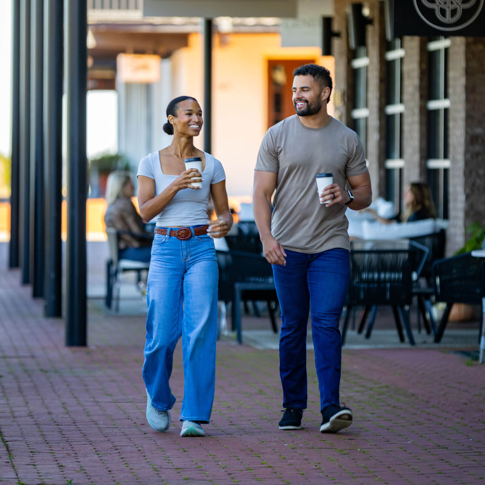 A man and woman walk and drink coffee on the sidewalk in front of the Town Center's businesses.